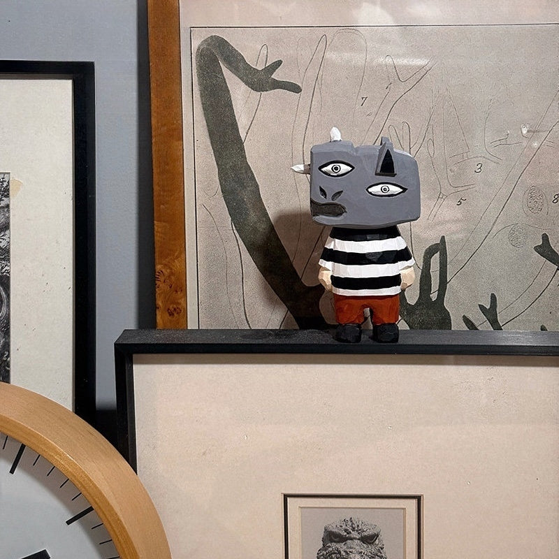 Guernica-inspired hand-carved wooden monster figure displayed on a shelf in front of abstract art prints and a wall clock.
