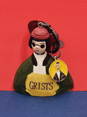 A hand-carved Mathilda movie keychain from Léon, wearing her signature olive green jacket and red hat, with a small charm showing her likeness, against a red background.
