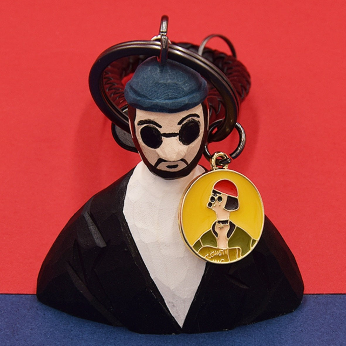 A close-up of a hand-carved Léon movie keychain against a red background, with a small circular charm depicting a stylized image of him.