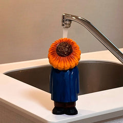 Hand-carved sunflower man placed under a running tap at a kitchen sink.