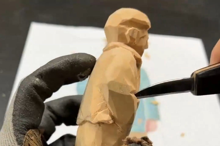 Close‑up of carving a wooden figurine, showing the knife shaping the sleeve and clothing folds — a detailed wood carving figures process for creating handcrafted wooden gifts.