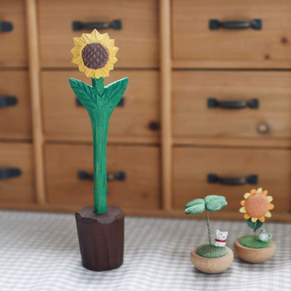 Carved sunflower pen standing in a brown wooden holder on checked fabric beside two small ornaments.