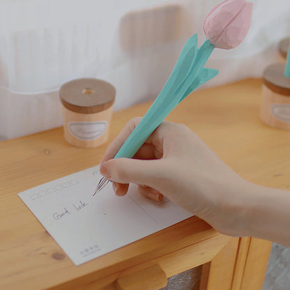 A hand writing on a postcard with a pink wooden tulip pen, with its pot holder nearby.