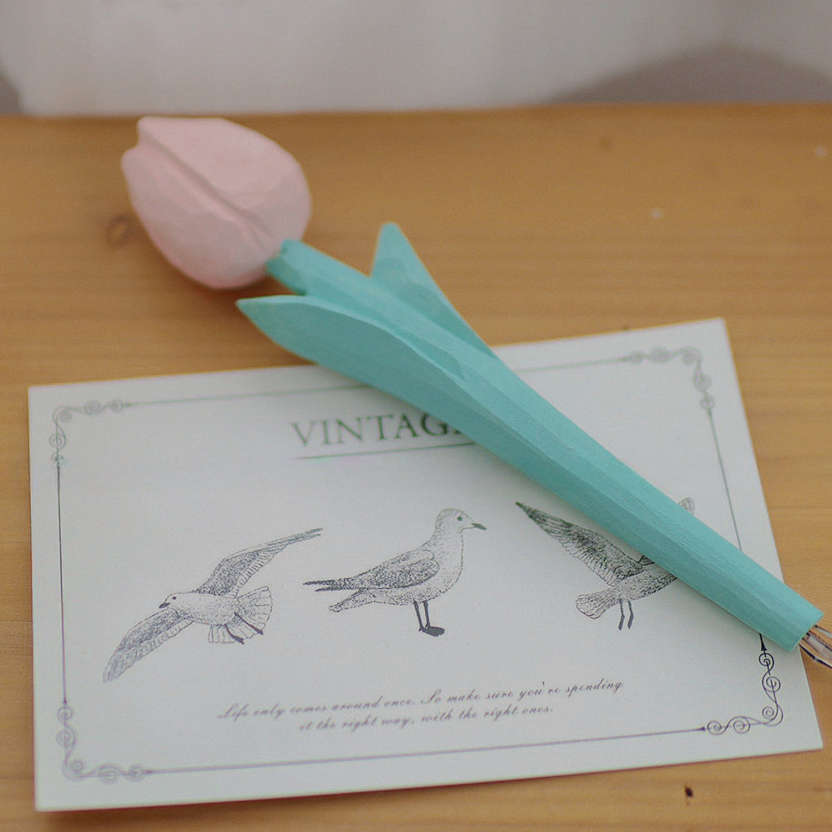 Pink wooden tulip pen resting diagonally across a bird postcard on a wooden desk.