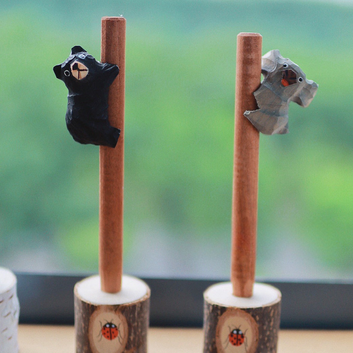 Close-up of wooden pens with hand-carved bear and koala figures in tree stump holders, set against a green landscape background.