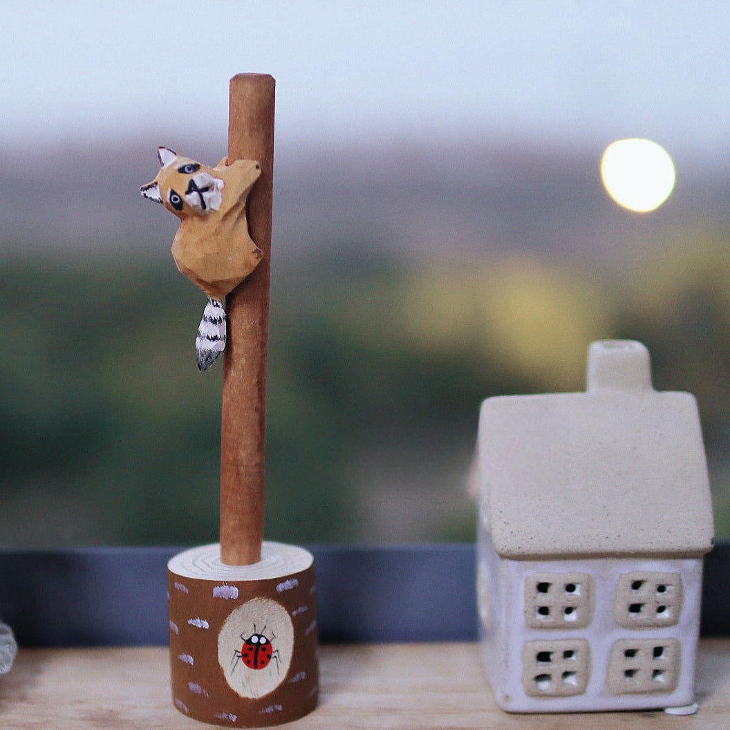 Wooden pen with a hand-carved raccoon figure in a tree stump holder, displayed with a scented candle and a ceramic house.