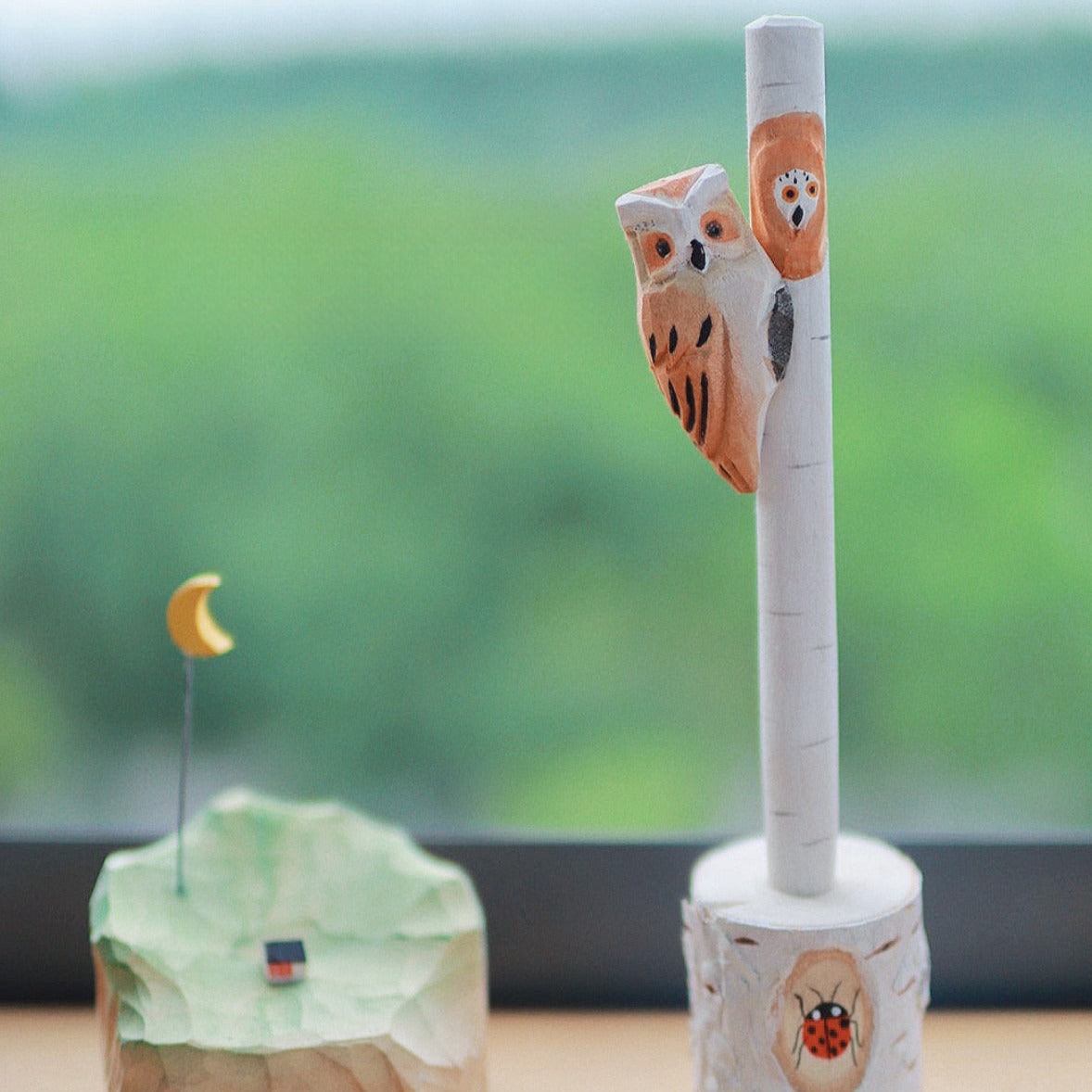 Wooden pen with a hand-carved owl figure in a tree stump holder, with a miniature moon and landscape decoration in the background.