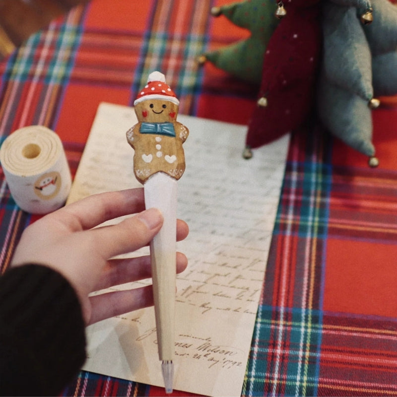 Hand holding a wooden gingerbread man pen with a red Christmas hat, with its pen holder and a fabric tree in the background