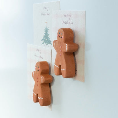 Two wooden gingerbread man magnets viewed from the side, showing their thick edges as they hold Christmas cards