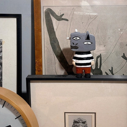 Guernica-inspired hand-carved wooden monster figure displayed on a shelf in front of abstract art prints and a wall clock.