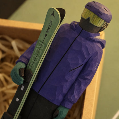 Angled close-up of a hand-painted wooden skier holding green skis, with carved jacket folds