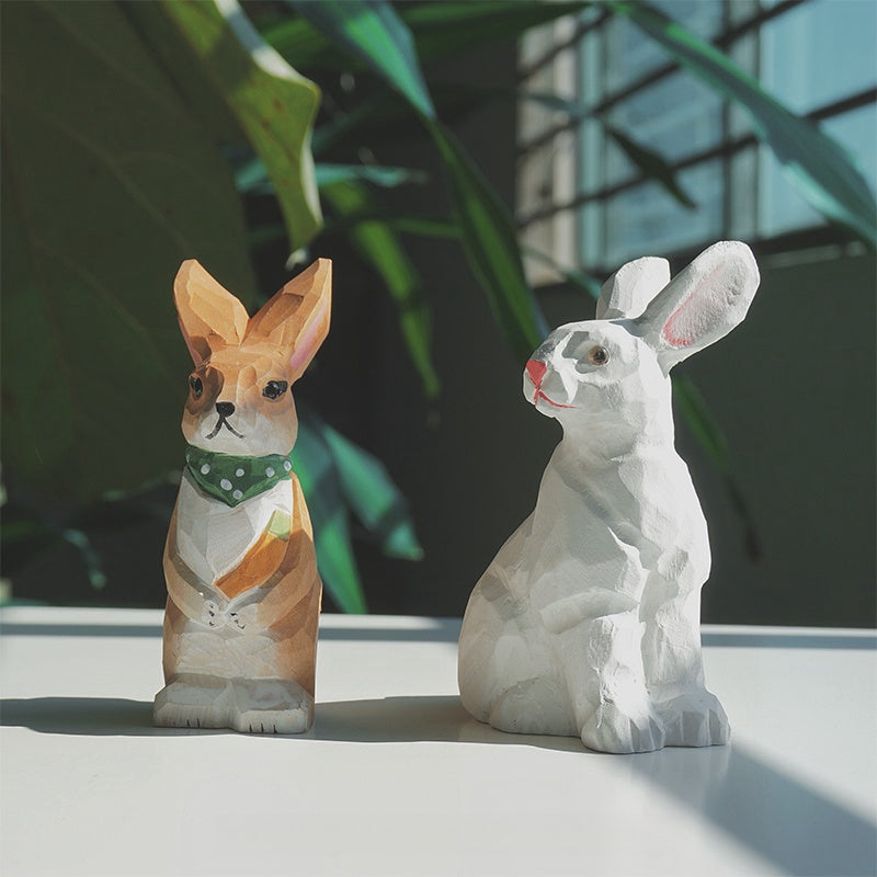 Two hand-carved bunny figurines on a sunlit tabletop, one brown-and-white with a green scarf and carrot and one seated white bunny