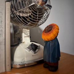 Sunflower head wooden figure standing next to a vintage fan and books on a shelf.
