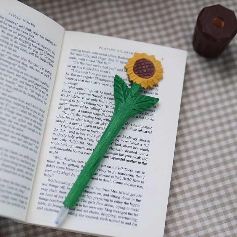 Wooden sunflower pen resting on an open book with its writing tip visible and brown pot holder nearby.