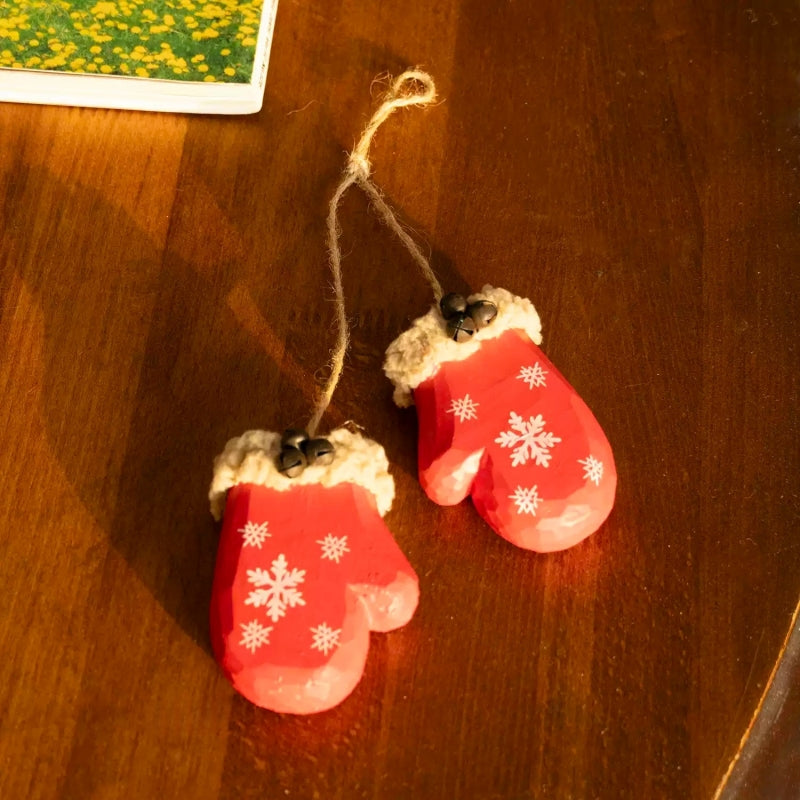 Two red wooden mitten ornaments and their connecting twine lying on a wooden table beside floral artwork