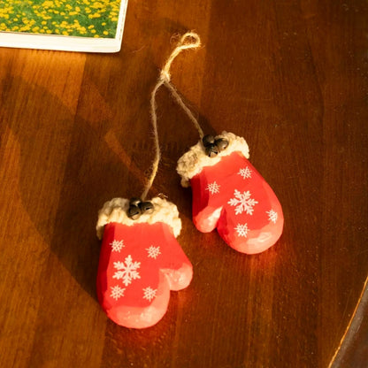 Two red wooden mitten ornaments and their connecting twine lying on a wooden table beside floral artwork