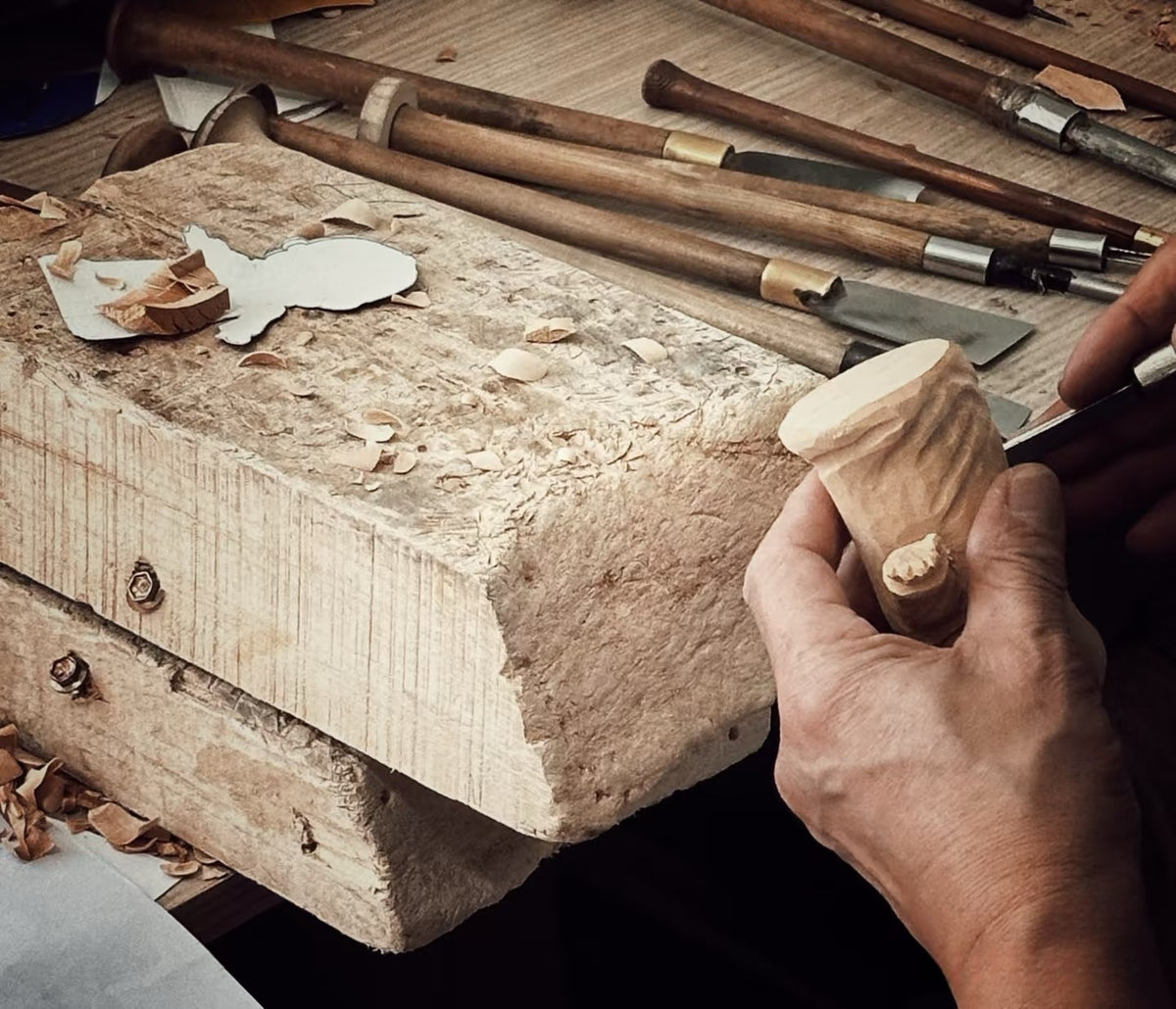 Hands carving a wooden form at a workbench with a row of carving tools