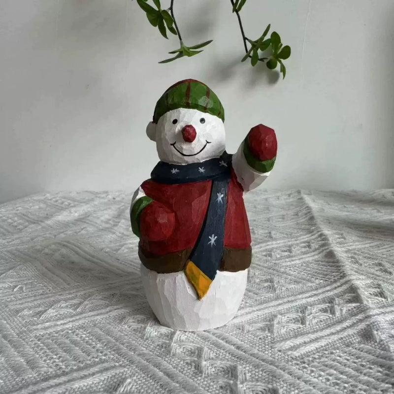 Wooden snowman figurine with a raised red mitten, navy scarf, and striped hat on textured white fabric