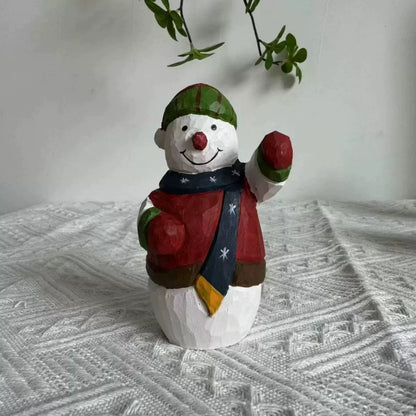 Wooden snowman figurine with a raised red mitten, navy scarf, and striped hat on textured white fabric